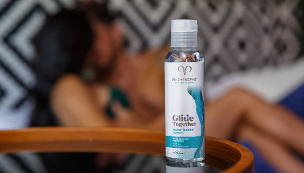 Promescent water lubricant on a glass table with a blurred out couple engaged in foreplay in the background