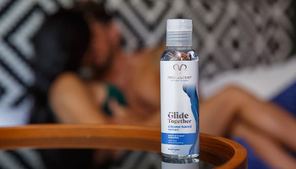 Promescent silicone lubricant on a glass table with a couple engaged in foreplay in the background