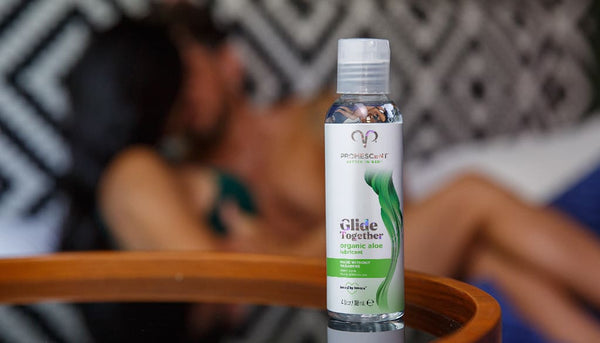Promescent organic aloe lubricant on a glass table with a couple in the background engaged in foreplay